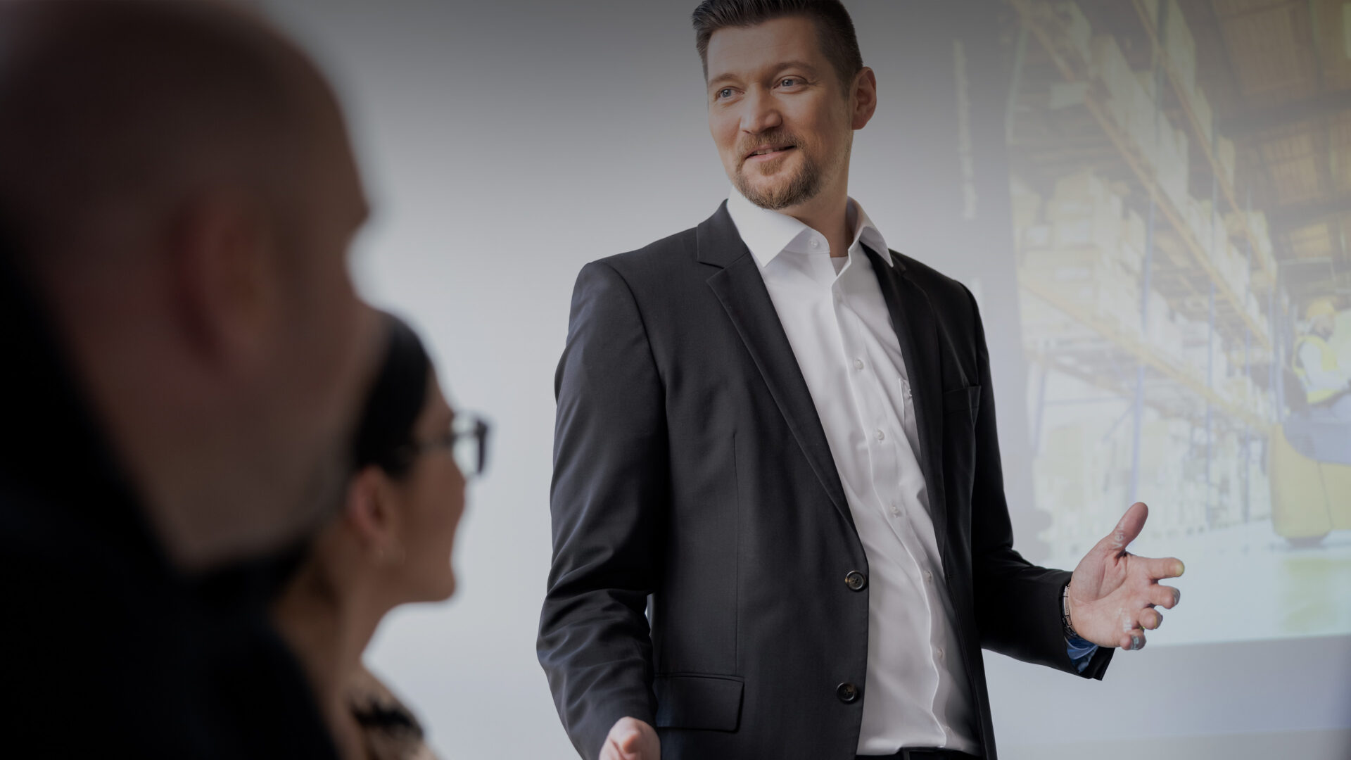 Ein Mann im Anzug steht vor einer projizierten Leinwand und spricht zu zwei sitzenden Personen während einer Geschäftspräsentation oder -sitzung.