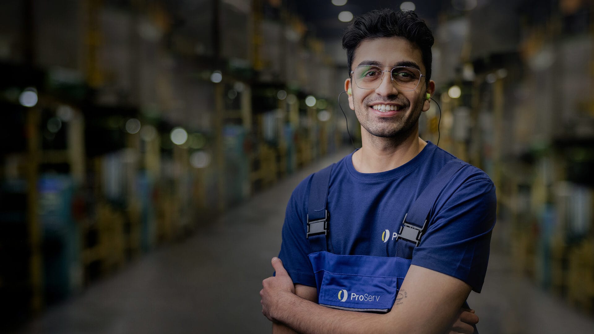 Ein lächelnder Mann mit Brille und blauer Arbeitsuniform steht mit verschränkten Armen in einem Lagerhaus. Im Hintergrund sind Regale und Industrieanlagen zu sehen, die leicht verschwommen sind.