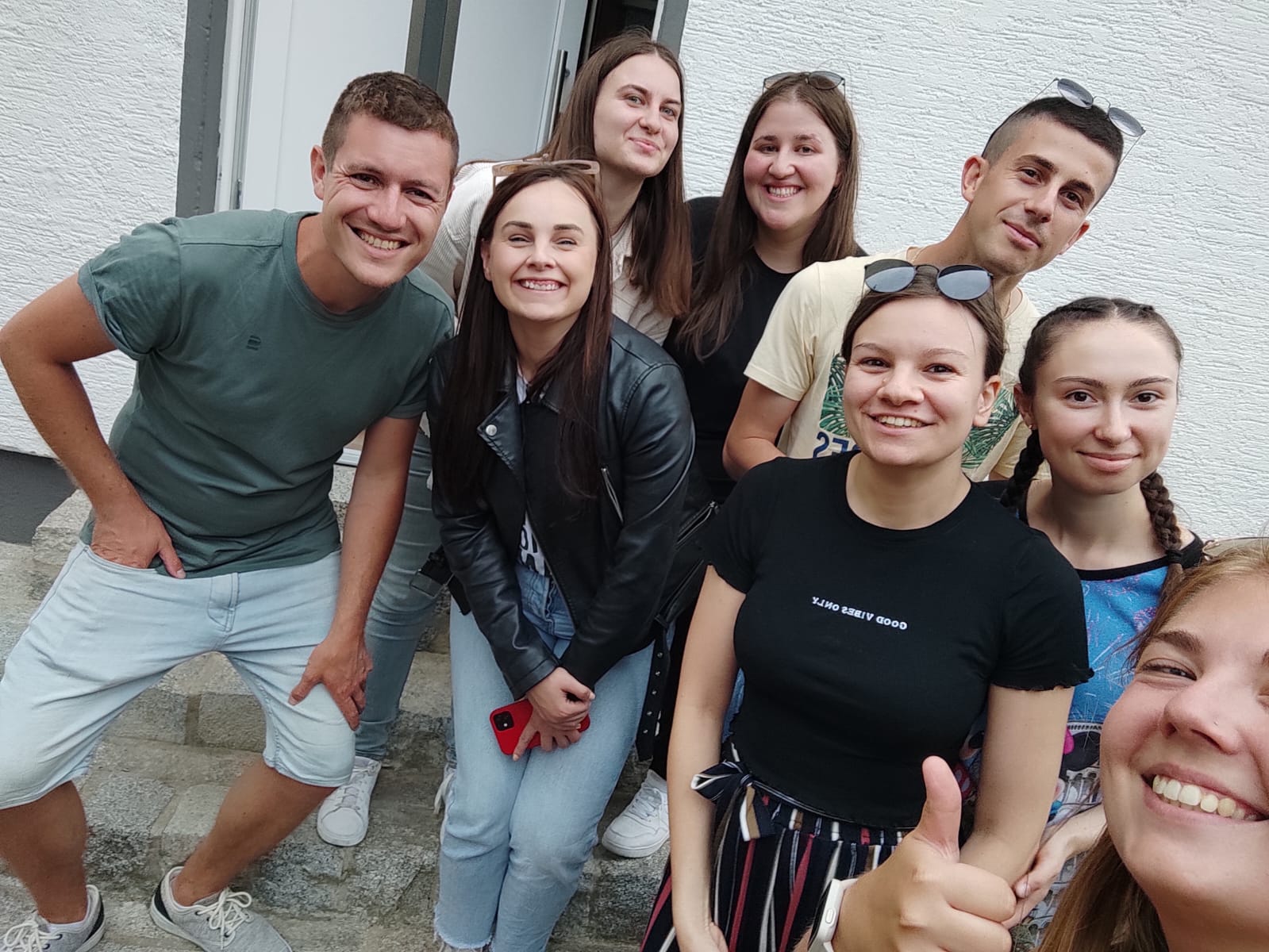 Eine Gruppe von acht lächelnden jungen Erwachsenen posiert gemeinsam für ein fröhliches Gruppen-Selfie im Freien.