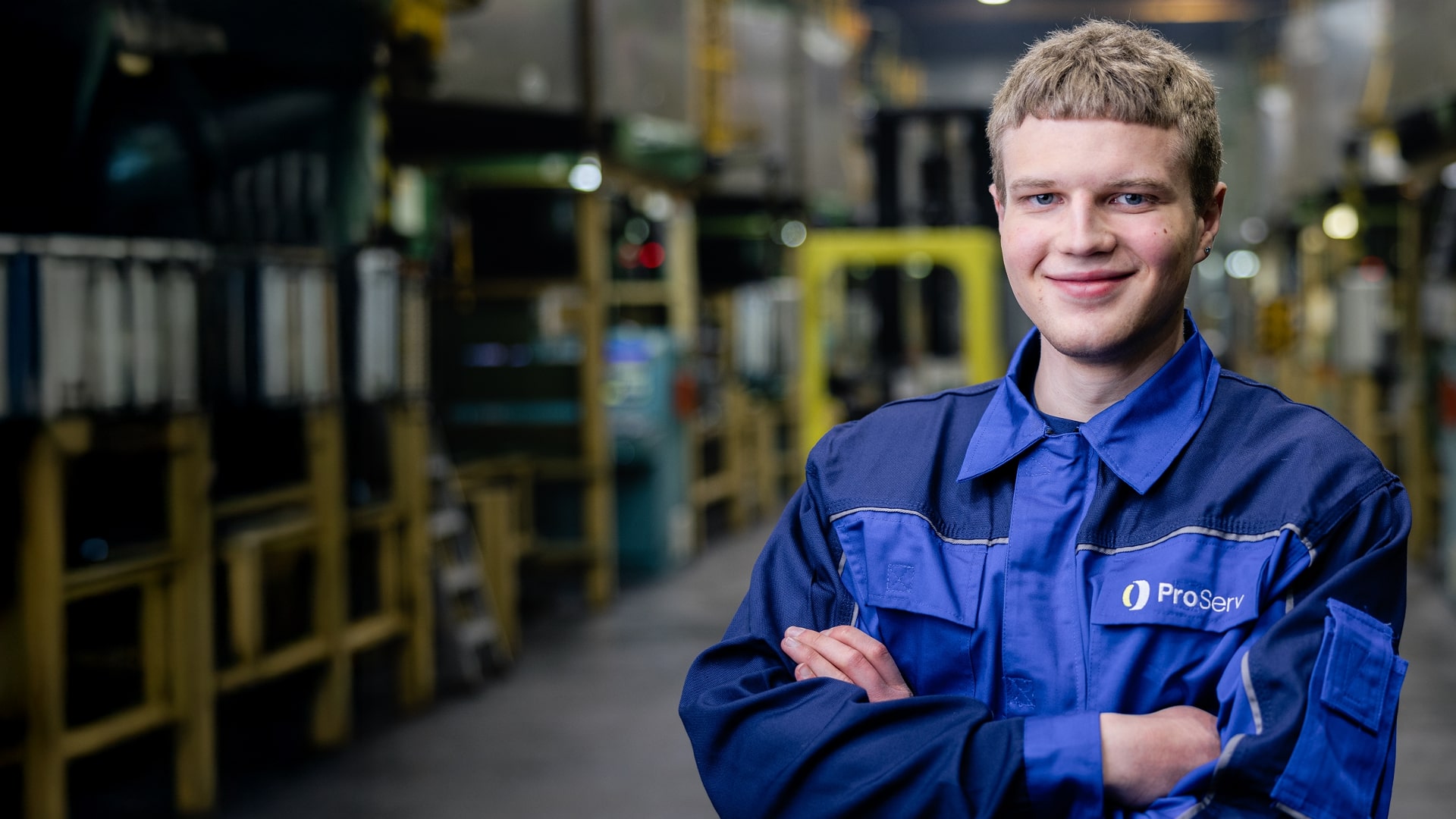 Junger Mann in blauer Uniform steht mit verschränkten Armen und lächelt in einer industriellen Fabrikumgebung.