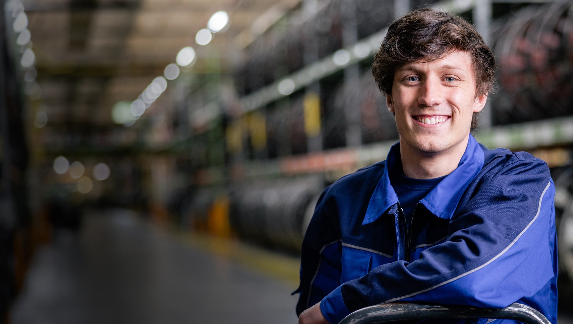Ein junger Mann in einer blauen Arbeitsuniform lächelt in einem großen Lagerhaus oder einer Industrieanlage.