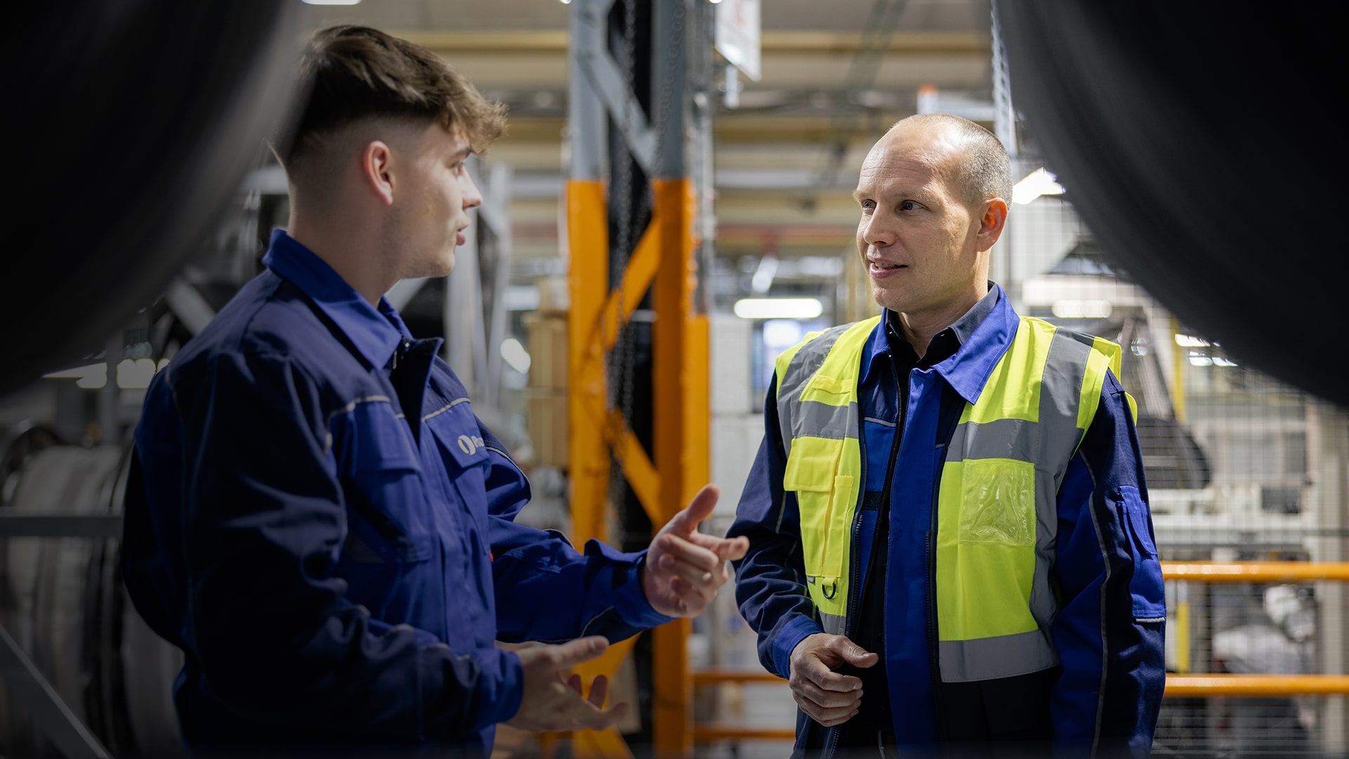 Zwei Männer in Arbeitsuniformen unterhalten sich in einer Industrieanlage, einer trägt eine gelbe Sicherheitsweste.